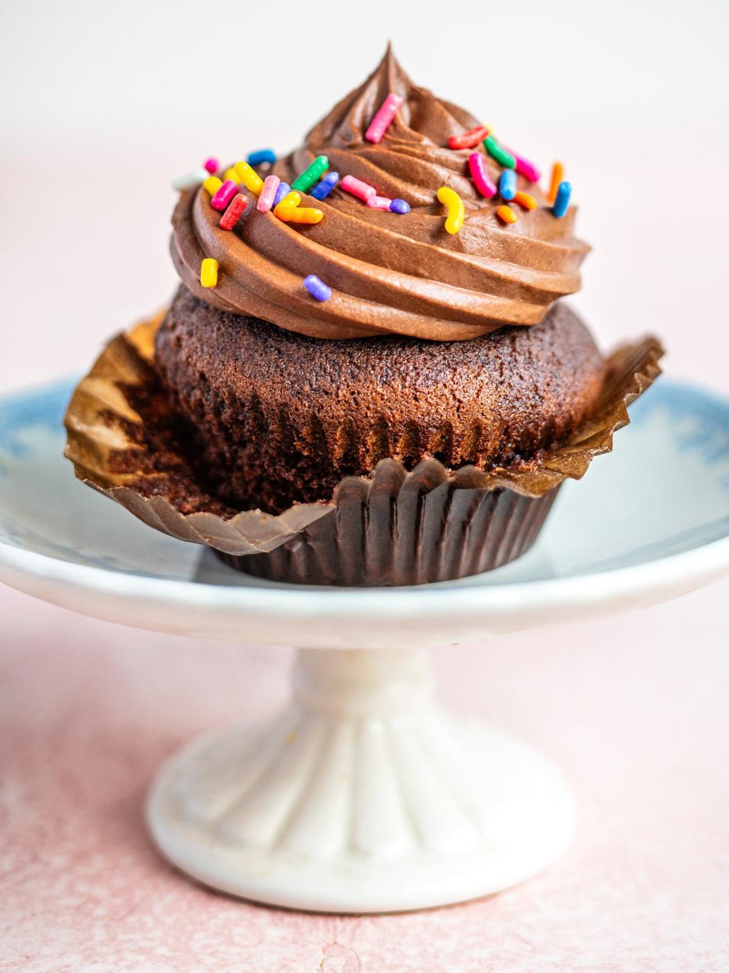 An air fryer chocolate cupcake with chocolate frosting and colorful sprinkles sits in a brown paper liner on a white cake stand with a blue rim.