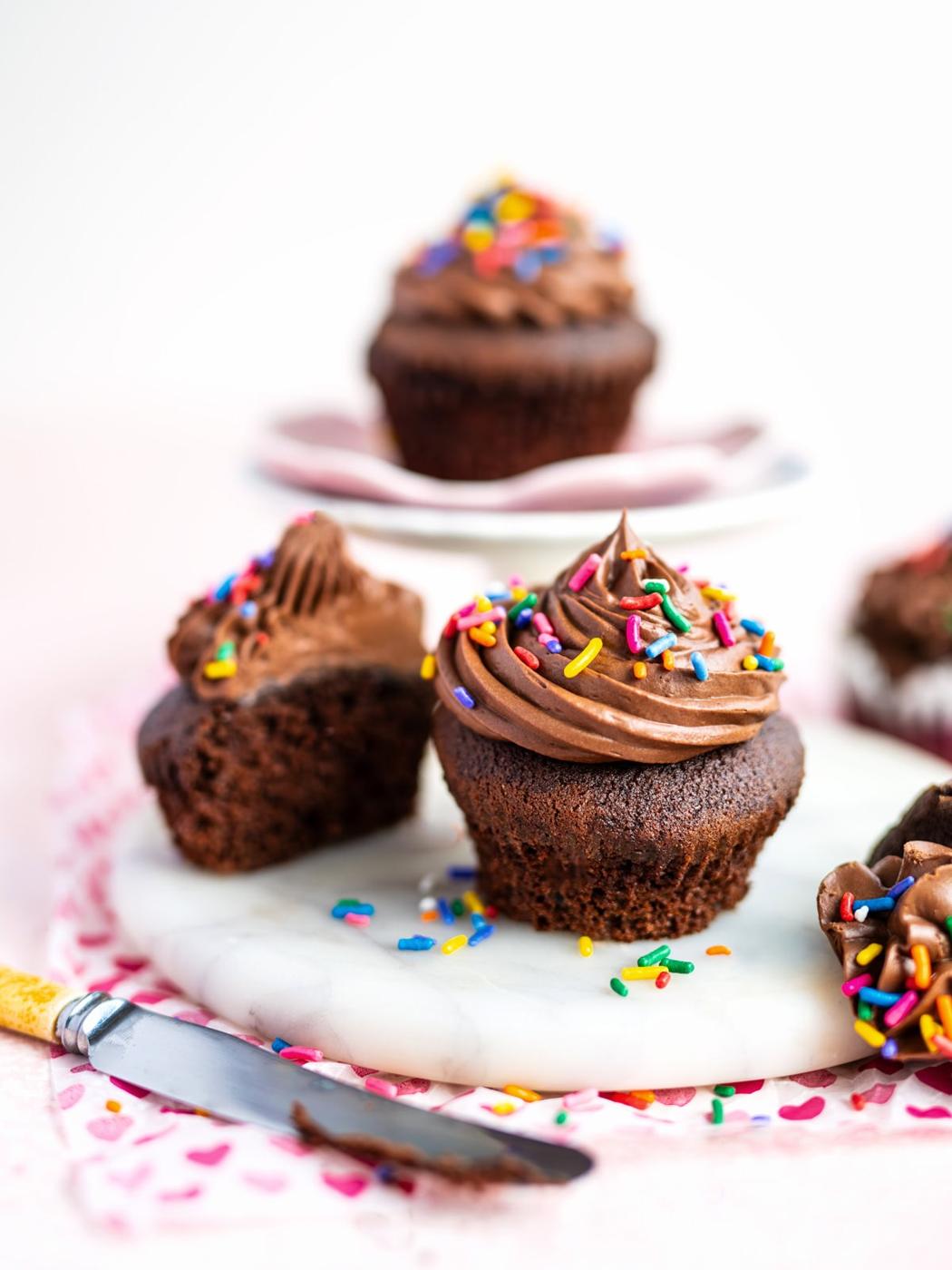 Air fryer chocolate cupcakes with chocolate frosting and rainbow sprinkles sit on a white marble surface; one cupcake is cut in half, showing its moist interior, with a yellow-handled knife nearby.
