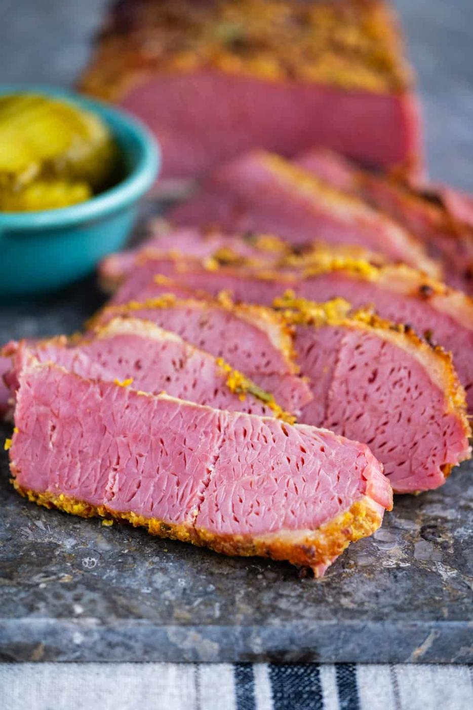 Sliced air fryer corned beef with a seasoned crust is arranged on a stone surface. A small bowl of pickles sits in the background, highlighting the pink, tender meat.