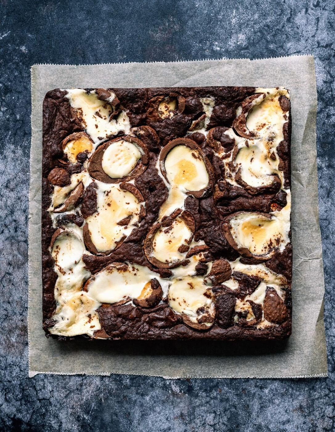 A square, baked chocolate brownie with swirls of cream cheese on top sits on parchment paper, placed on a textured dark surface—a tempting treat inspired by Creme Egg Brownies in your air fryer.