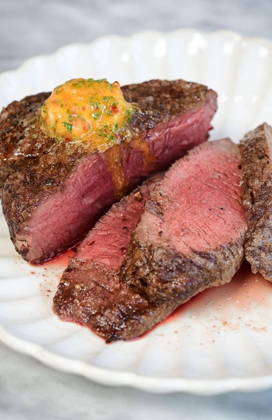 A sliced medium-rare steak on a white plate, topped with a dollop of herbed compound butter that is melting over the meat.