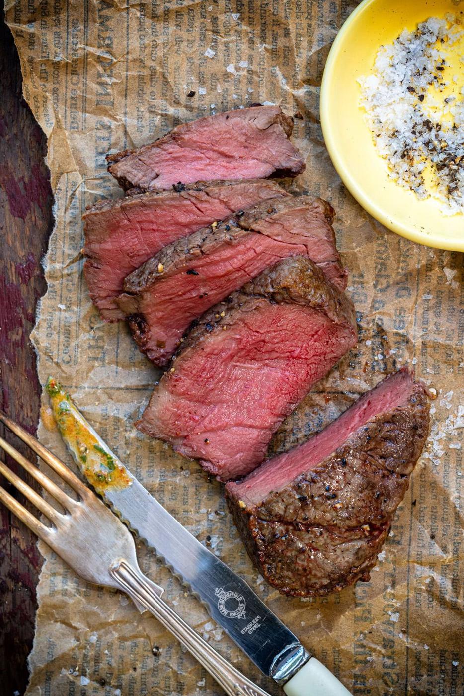 Sliced medium-rare steak on brown parchment with a vintage fork and knife, next to a yellow dish of salt and pepper. The steak is juicy and pink inside with a seared, peppered crust.