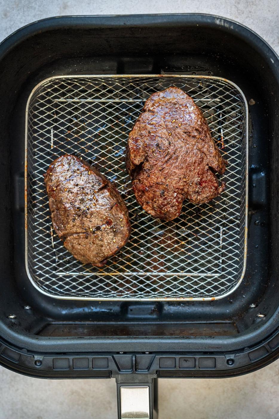 Two cooked fillet steaks resting on the metal rack inside an air fryer basket.