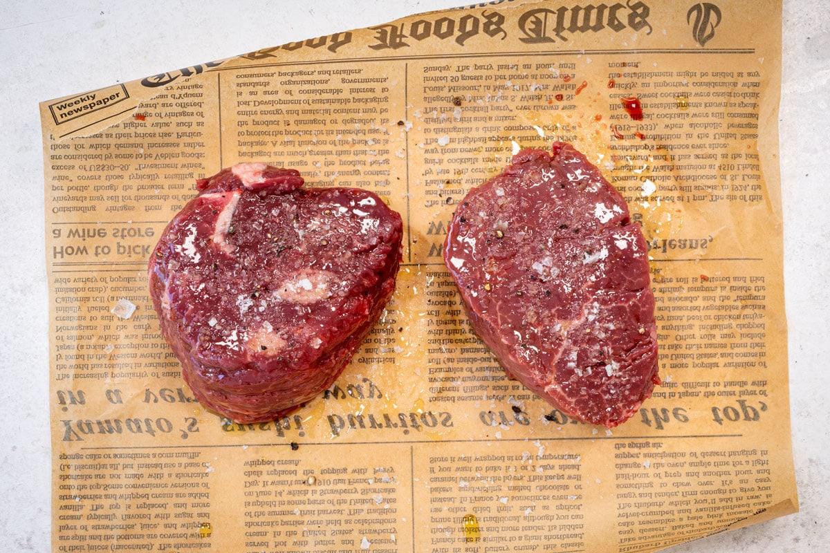 Two raw beef steaks seasoned with salt and pepper sit on a piece of newspaper on a light-colored surface.