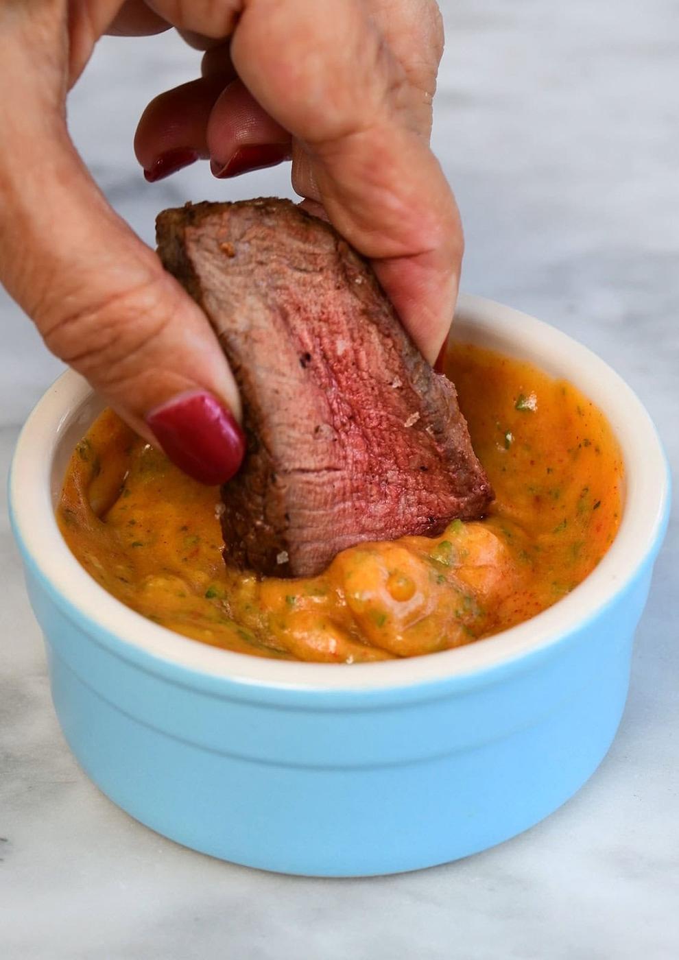 A hand with red nail polish dips a slice of cooked steak into a cowboy butter dip in a small blue ramekin.