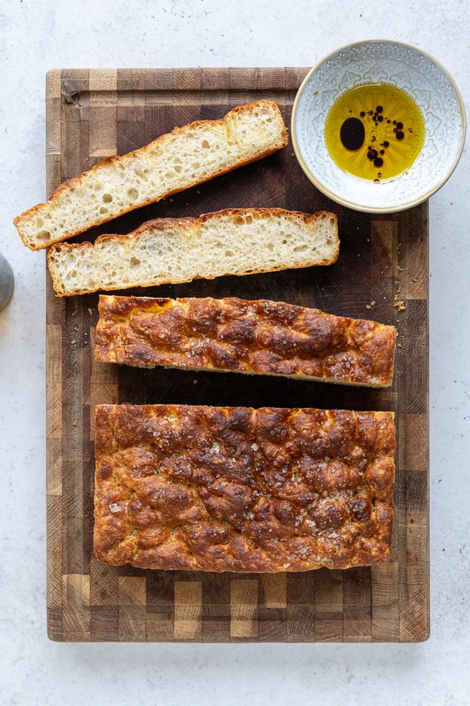 A fluffy loaf of Air Fryer Focaccia bread rests on a wooden cutting board, with two slices cut and placed above it. A small bowl of olive oil with herbs sits next to the bread.