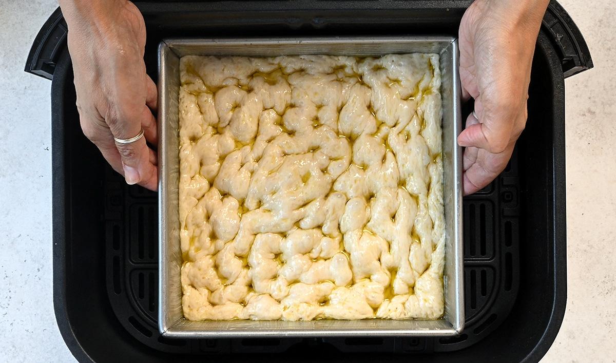 Hands placing a square pan of unbaked, dimpled focaccia dough drizzled with olive oil into an air fryer—an easy focaccia recipe for delicious Air Fryer Focaccia bread.