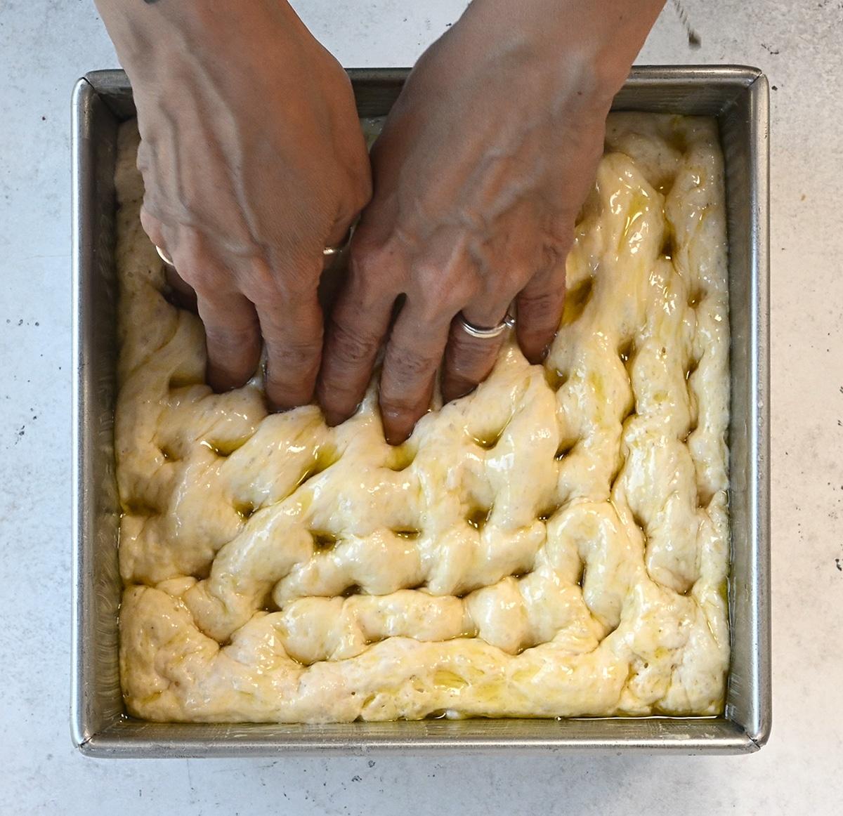 Two hands press finger indentations into glossy, oil-brushed focaccia dough in a square metal pan, preparing this fluffy, easy focaccia recipe for baking. Perfect for making Air Fryer Focaccia at home.