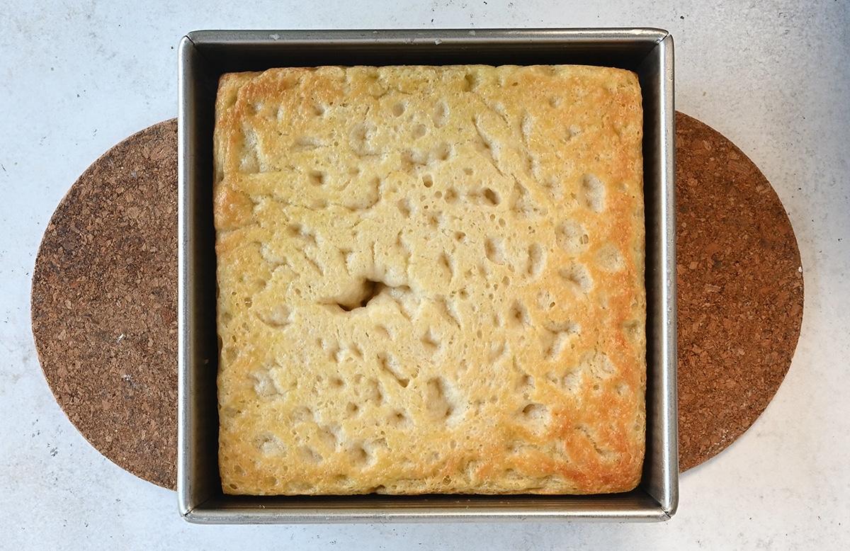 A square loaf of pillowy, golden-brown bread with a slightly uneven, airy surface sits in a metal baking pan on a round cork mat—perfect Air Fryer Focaccia.