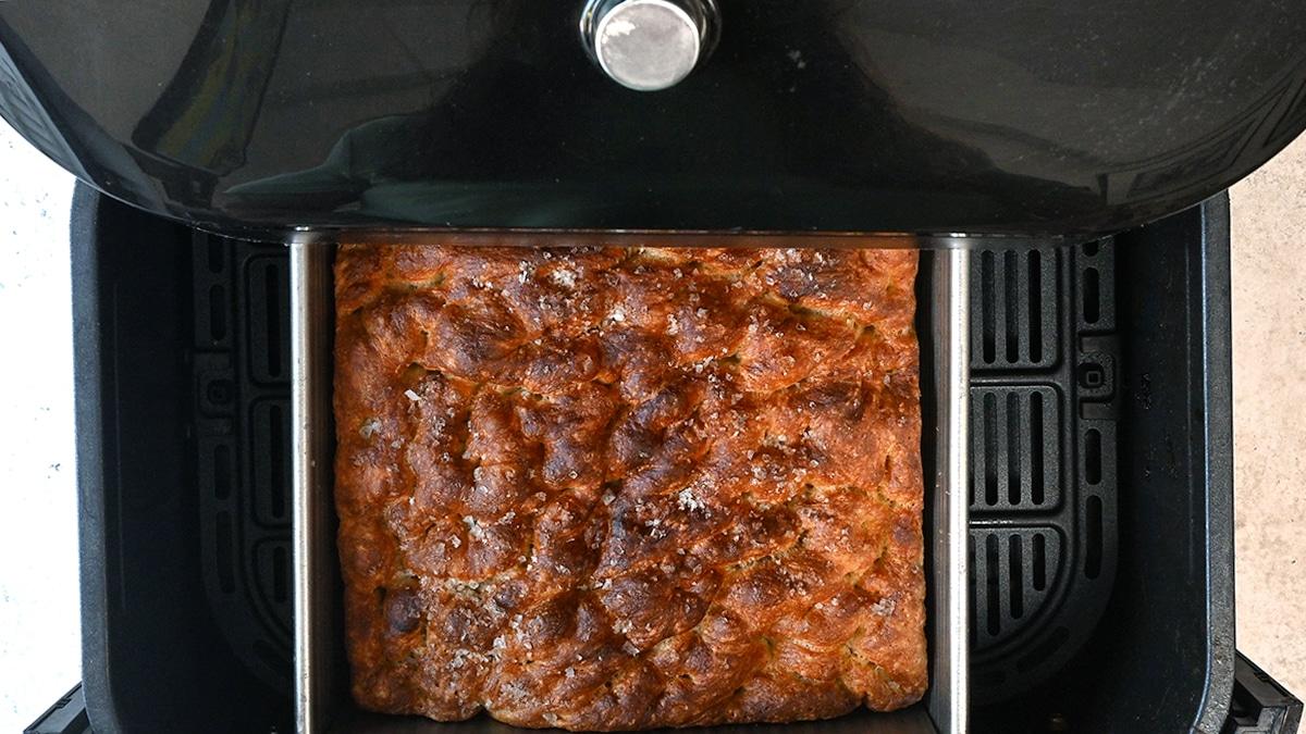 A golden-brown Air Fryer Focaccia sits in a rectangular pan inside an open air fryer, viewed from above. The bread looks fluffy and crisp with a beautifully textured, dimpled top—an easy focaccia recipe for any home baker.