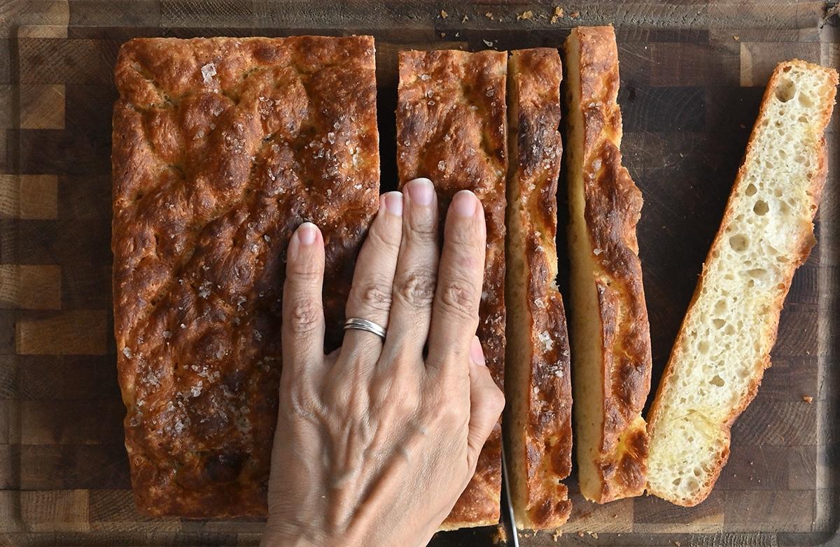 A hand with a silver ring slices a loaf of golden-brown focaccia bread on a dark wooden cutting board. The bread has a crisp crust and pillowy, airy texture inside—perfect for those seeking an easy focaccia recipe.