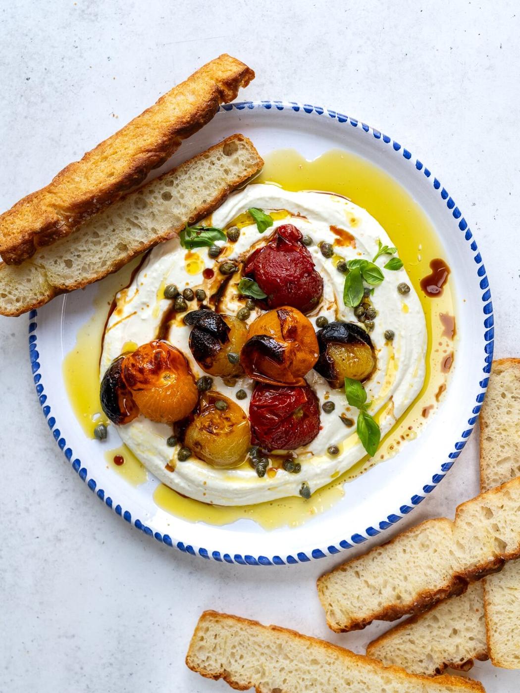 A plate with fluffy whipped cheese topped with roasted cherry tomatoes, olives, capers, fresh basil, and olive oil, served with slices of toasted bread on a white surface.