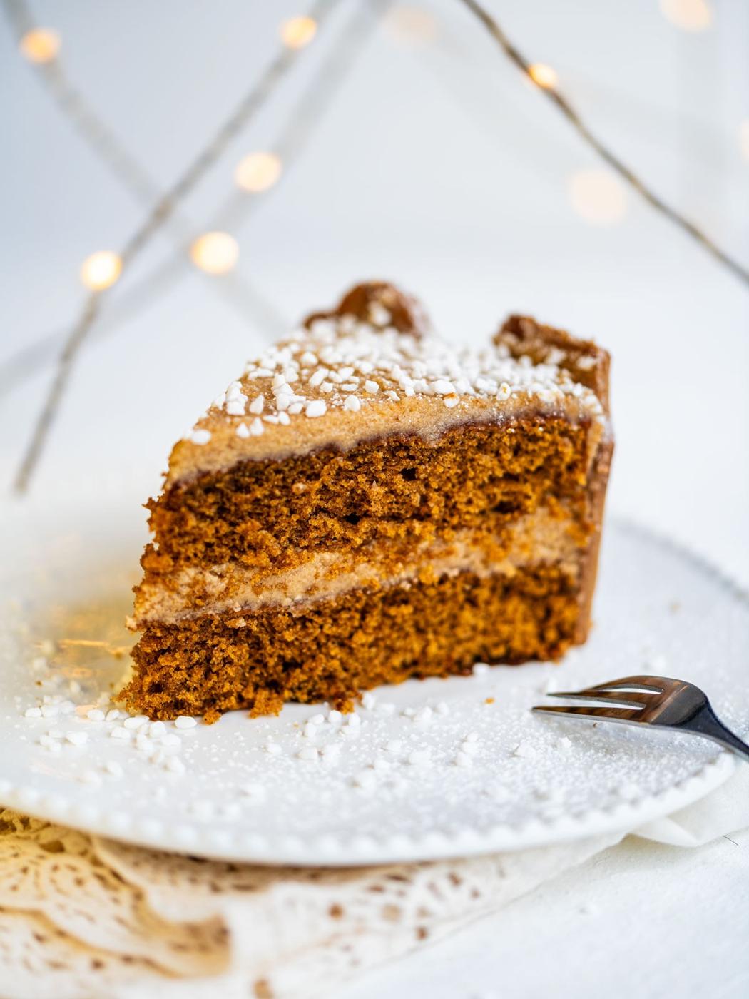 A slice of layered gingerbread cake with ermine frosting and sprinkled with white sugar crystals sits on a white plate, with a fork nearby and warm fairy lights in the blurred background.