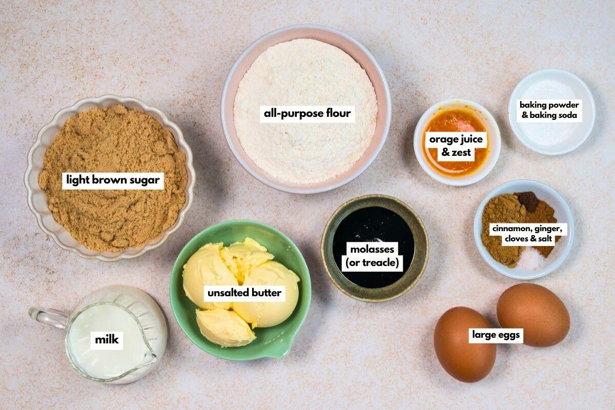 Top-down view of baking ingredients for Air Fryer Gingerbread Cake in bowls: light brown sugar, all-purpose flour, orange juice and zest, baking powder and soda, spices with salt, milk, unsalted butter, molasses (treacle), and two large eggs.
