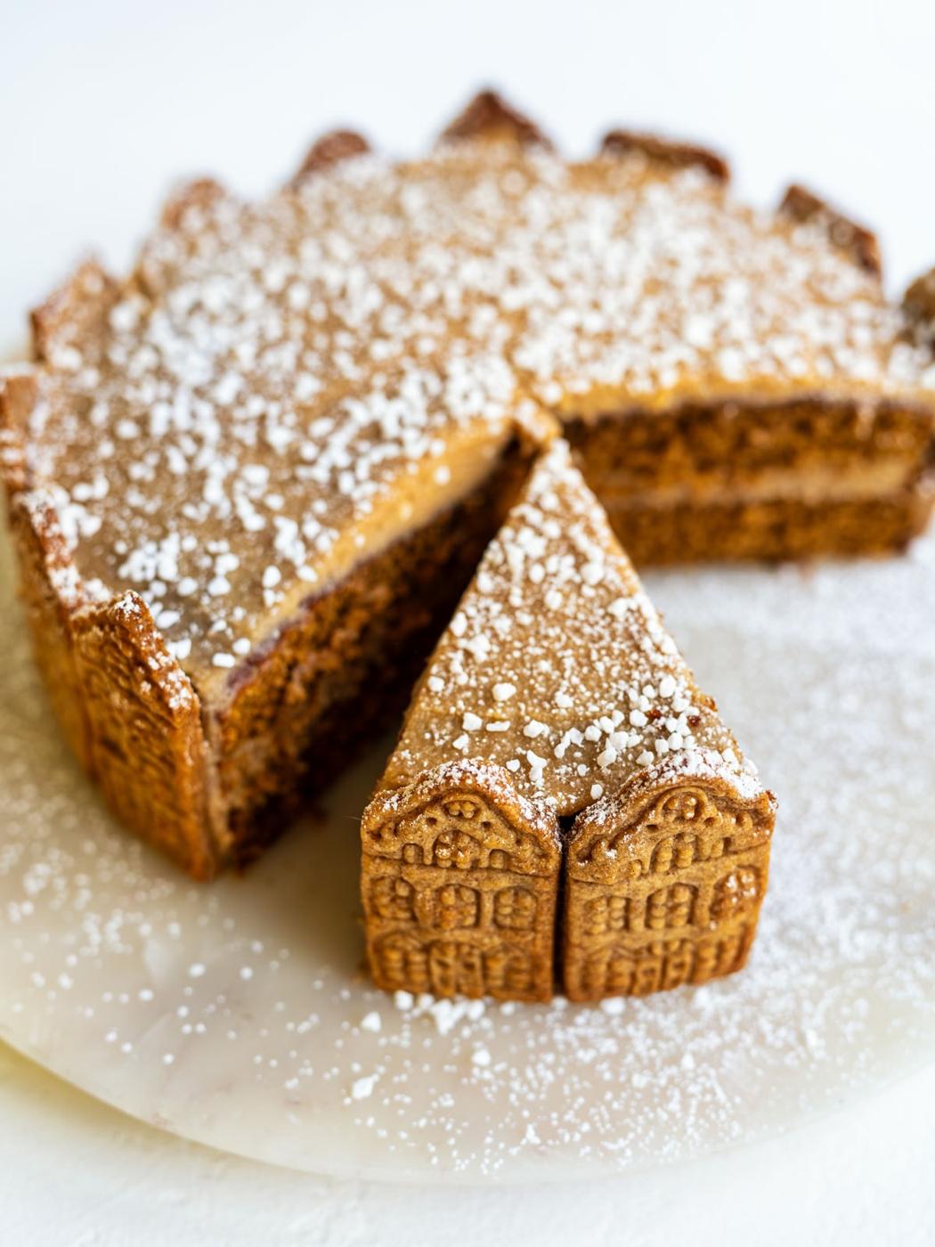 A round layered cake decorated with rectangular cookies around the sides, topped with powdered sugar. A single slice has been removed and is placed in front, showing the cookie detail.
