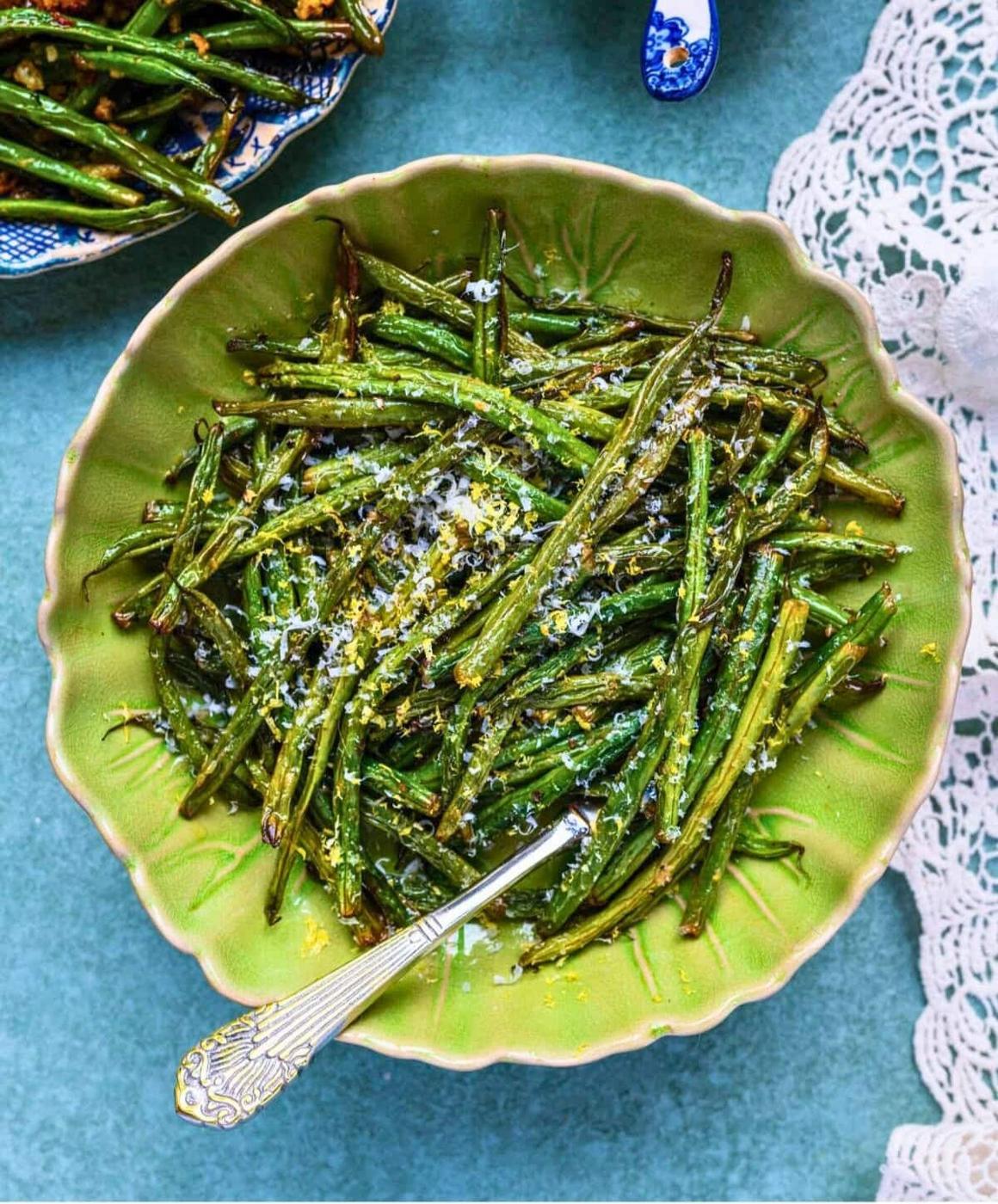 A green bowl filled with roasted green beans topped with grated cheese and lemon zest, with a silver fork resting inside. The bowl is set on a blue surface next to a lace cloth.