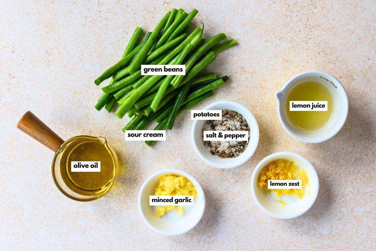 Labeled ingredients on a countertop: green beans, sour cream, olive oil, minced garlic, lemon zest, lemon juice, chopped potatoes, and a bowl of salt and pepper.