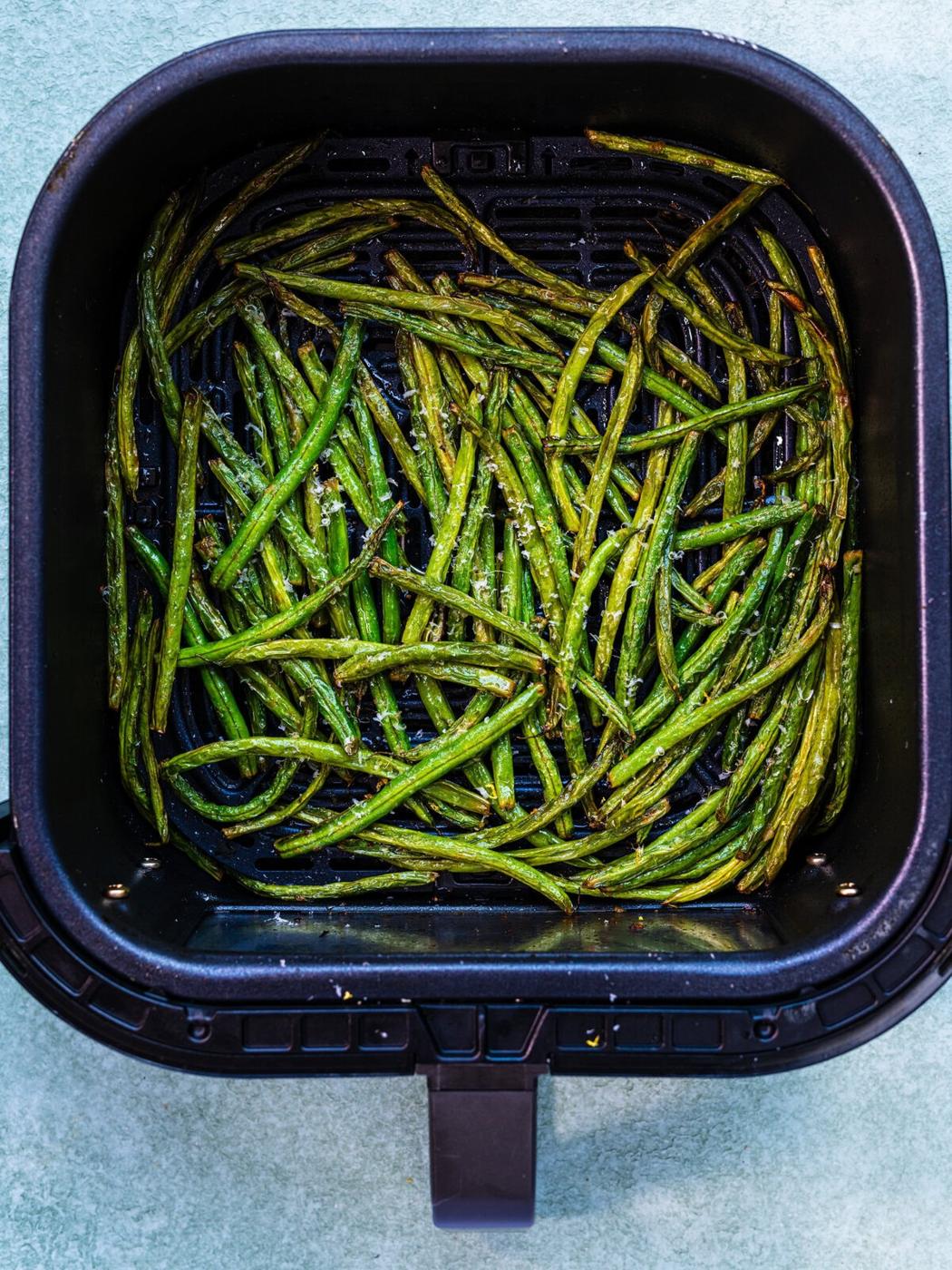 Green beans cooked in an air fryer basket, lightly browned and seasoned, sitting on a light-colored surface.