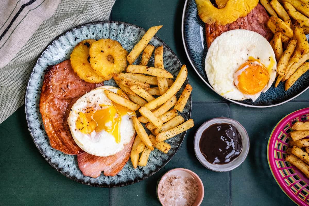 Two plates featuring air fryer ham steak, pineapple rings, fried eggs, and seasoned fries. A small bowl of salt sits next to a dish of dark sauce. A striped napkin is partly visible in the top left corner.