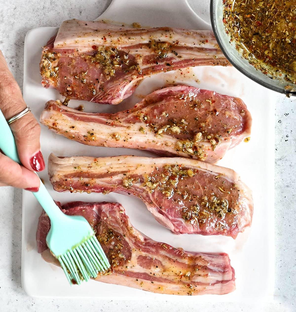 Four uncooked lamb chops on a white tray are being brushed with a herb and spice marinade using a silicone brush. A bowl of marinade sits nearby, and a hand is holding the brush.