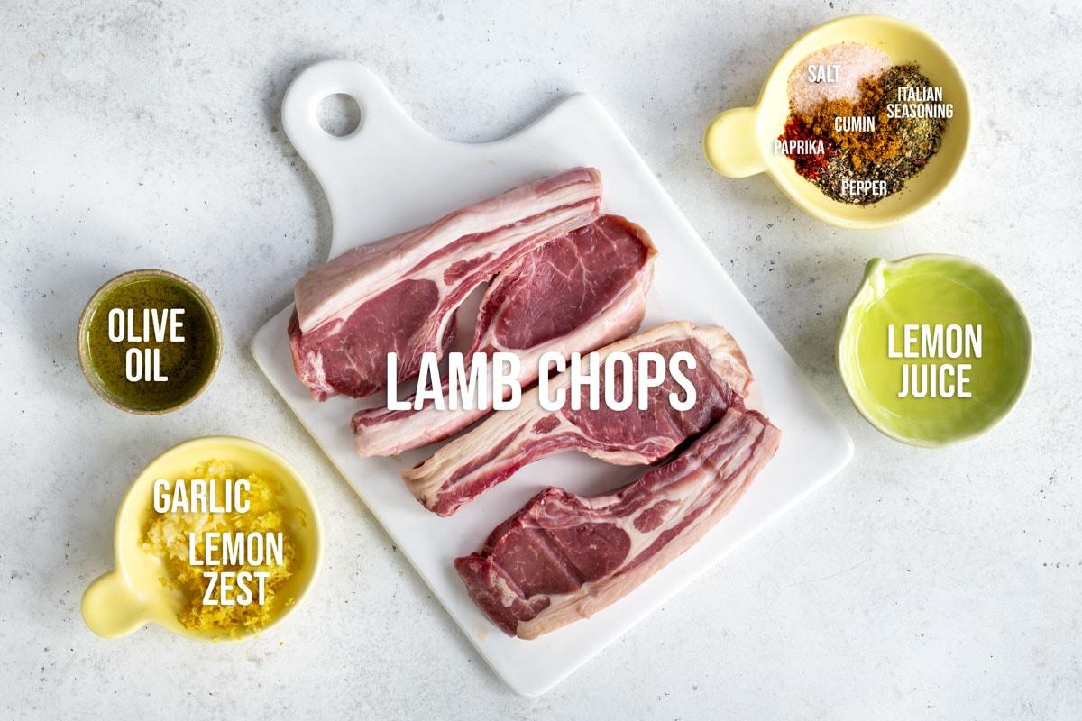Raw lamb chops on a white cutting board, surrounded by small bowls containing olive oil, garlic with lemon zest, lemon juice, and a mix of spices including salt, cumin, paprika, pepper, and Italian seasoning.