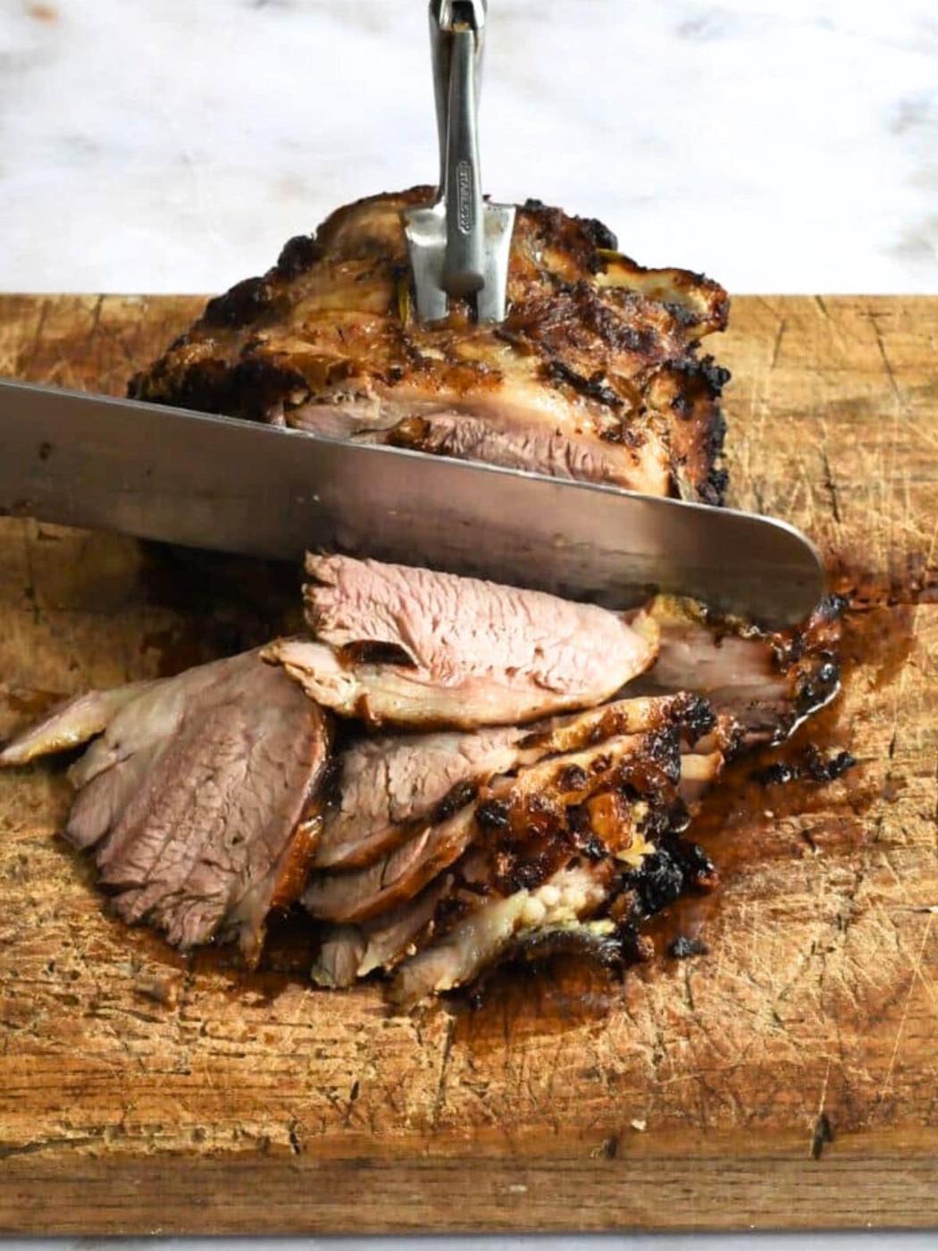 A knife slices through a cooked, browned Air Fryer Lamb Shoulder on a wooden cutting board, with several juicy slices fanned out and a meat fork holding the roast steady.