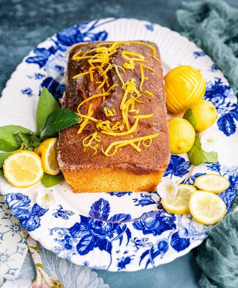 A rectangular air fryer lemon drizzle cake topped with lemon zest sits on a blue and white floral platter, surrounded by whole and sliced lemons, green leaves, and white flowers. A decorative cake server is nearby.