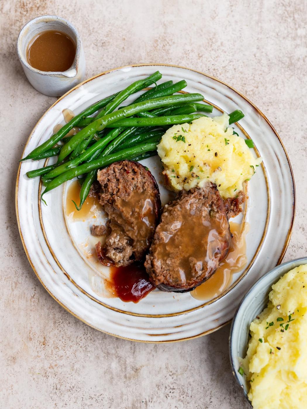 A plate with two slices of air fryer meatloaf topped with brown gravy, mashed potatoes with herbs, and green beans. Nearby are a small jug of gravy and a bowl of extra mashed potatoes.