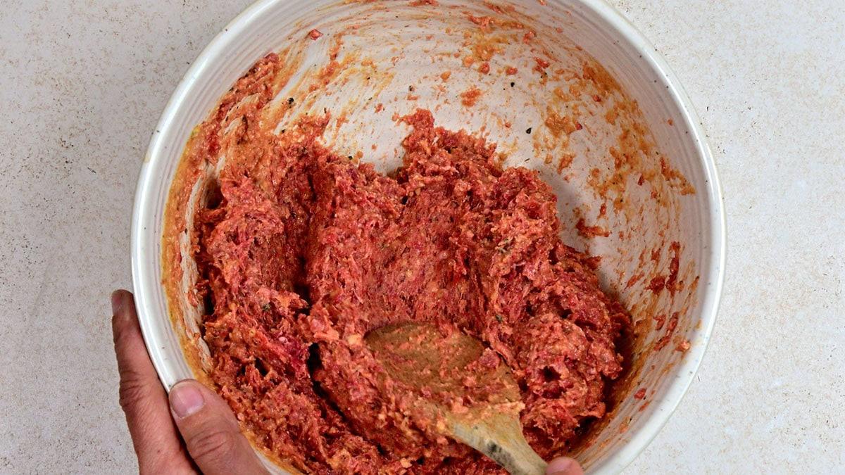 A person is mixing raw ground meat with seasonings in a white bowl using a wooden spoon. The mixture appears to be for making meatballs or meatloaf.