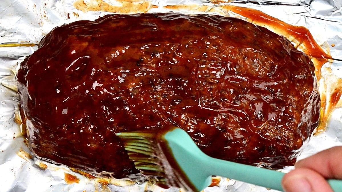 A hand uses a silicone brush to spread sauce over a cooked meatloaf resting on a sheet of aluminum foil.