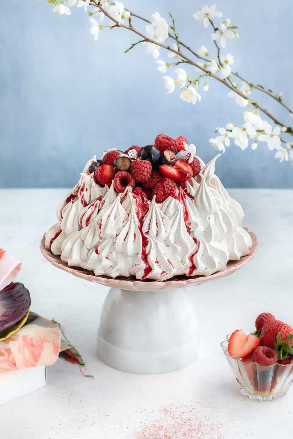 A beautiful Air Fryer Pavlova topped with fresh berries and berry sauce sits on a cake stand. Blossoming branches hang above, with a glass bowl of strawberries nearby. The pastel blue background adds a soft, inviting touch.