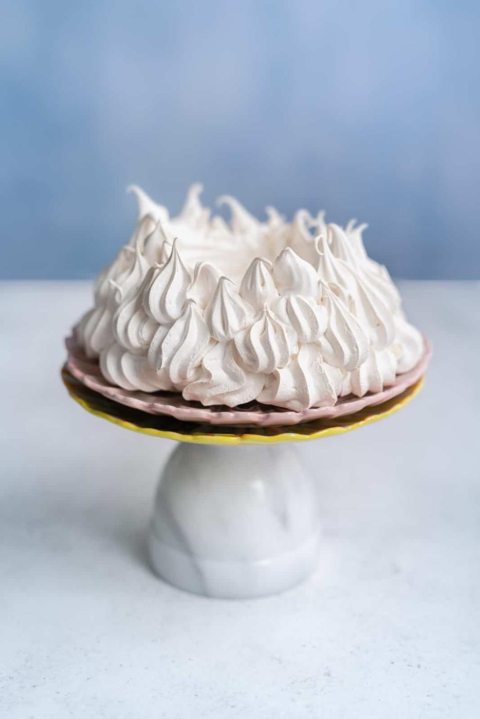 A meringue pie with golden peaks, reminiscent of an Air Fryer Pavlova, sits on a pink and yellow cake stand with a marble base, against a light blue background.