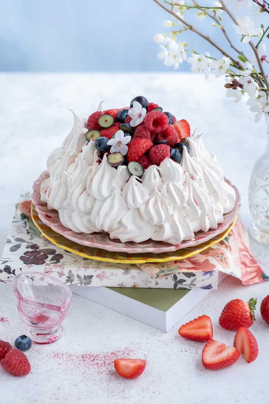 An Air Fryer Pavlova topped with fresh berries sits on patterned plates and books, surrounded by strawberries, blueberries, and a small glass. A vase of white blossoms is placed beside the beautiful dessert scene.