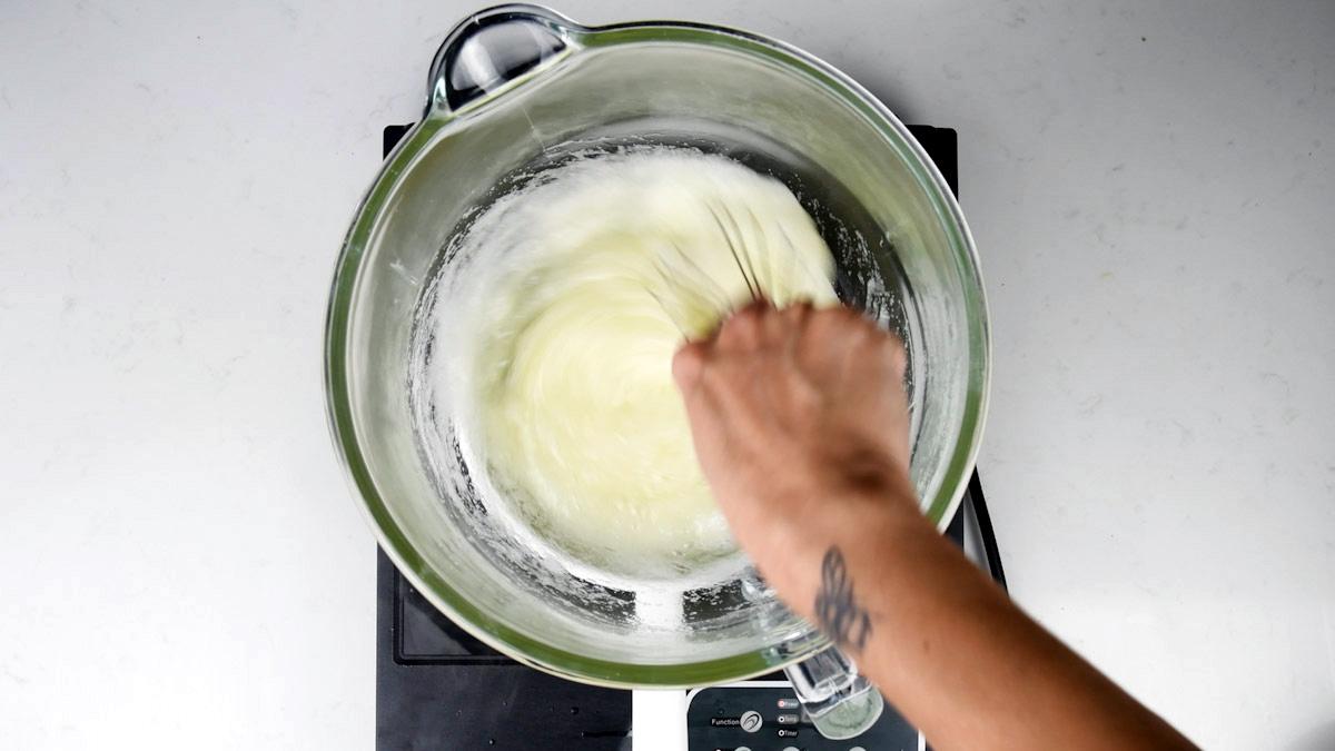 A person with a butterfly tattoo on their wrist is whisking a white mixture for Meringue Kisses in a glass bowl placed on a black stovetop.