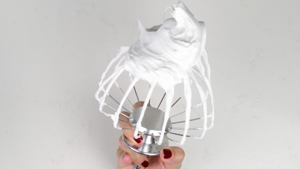 A hand holds a metal mixer whisk covered in thick, glossy meringue—perfect for making Meringue Kisses Cookies—against a white background.