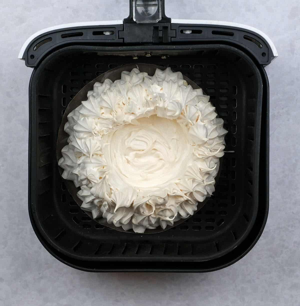 A round Air Fryer Pavlova with piped edges sits inside the basket, viewed from above. The meringue is white and features a swirled, decorative border.