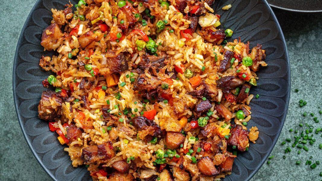 Three dark bowls filled with colorful fried rice, featuring diced vegetables, green peas, and crispy pieces of roast pork, are set on a gray surface, with chopped herbs sprinkled nearby.