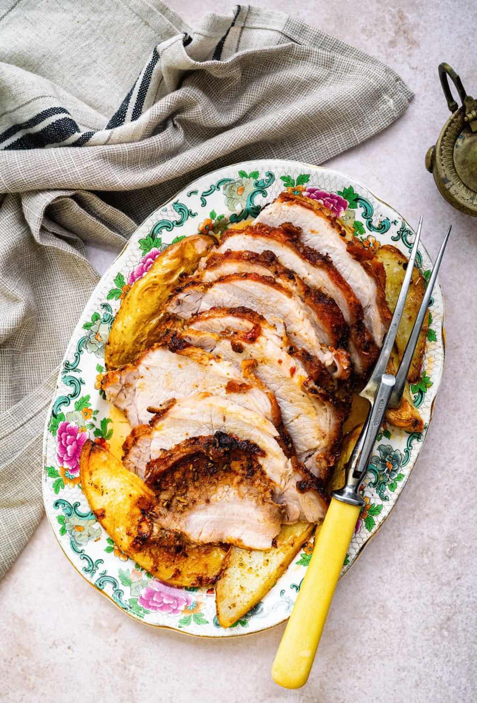 A platter with sliced roast pork and golden potato wedges, reminiscent of an Air Fryer Prime Rib feast, served on a floral-patterned dish with a yellow-handled carving fork, next to a folded cloth napkin.