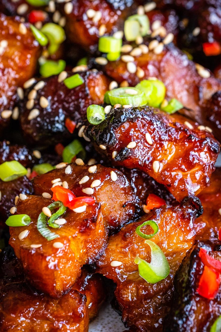 Close-up of air fryer pork belly bites, glazed and caramelized, topped with sliced green onions, sesame seeds, and bits of red chili, showcasing a shiny, sticky texture and vibrant garnishes.