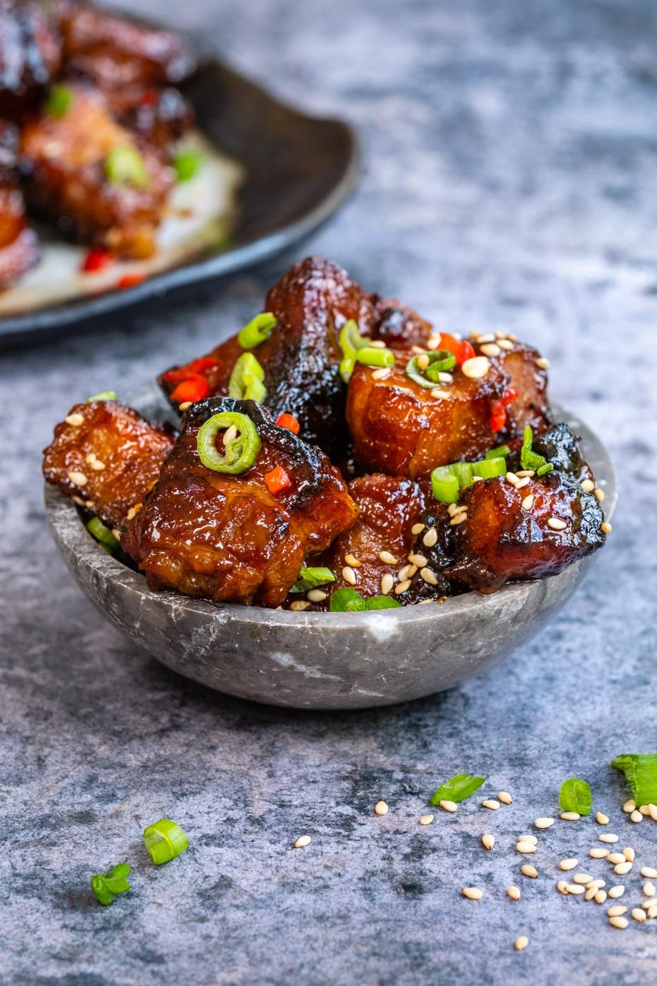 A small bowl filled with glazed, caramelized Air Fryer Pork Belly Bites topped with sliced green onions, red chili, and sesame seeds, with more pork belly bites air fryer style displayed on a plate in the background.