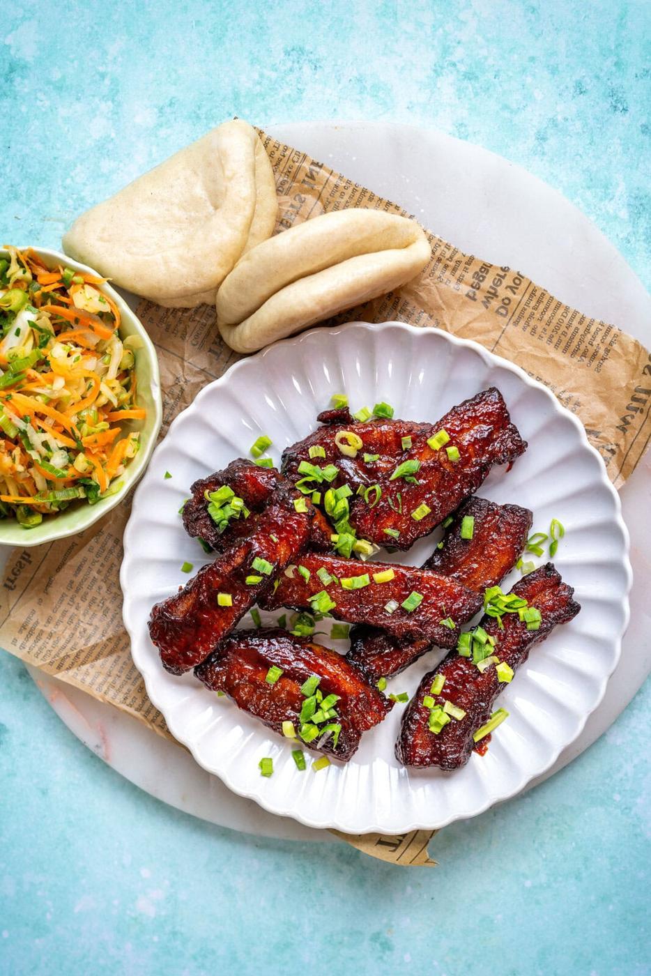 A white plate with glazed pork slices topped with chopped green onions, served next to a bowl of vegetable slaw and two folded steamed buns on a piece of newspaper over a marble surface.