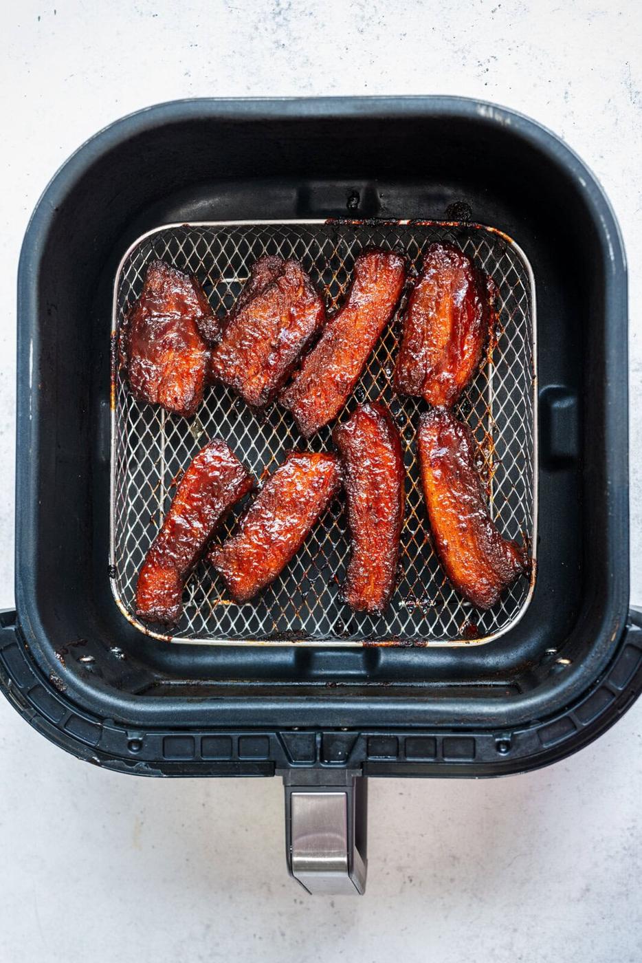 Eight pieces of glazed, cooked pork belly slices are arranged inside the basket of an air fryer on a light surface. The pork belly appears caramelized and crispy on the edges.