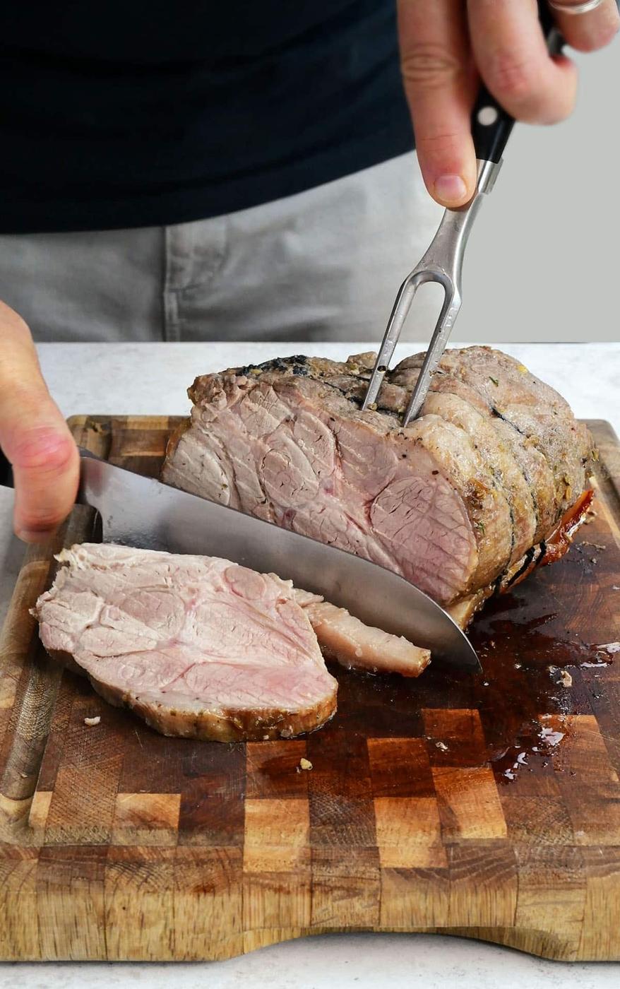 A person slices a juicy, tender roast pork loin on a wooden cutting board using a large knife and fork