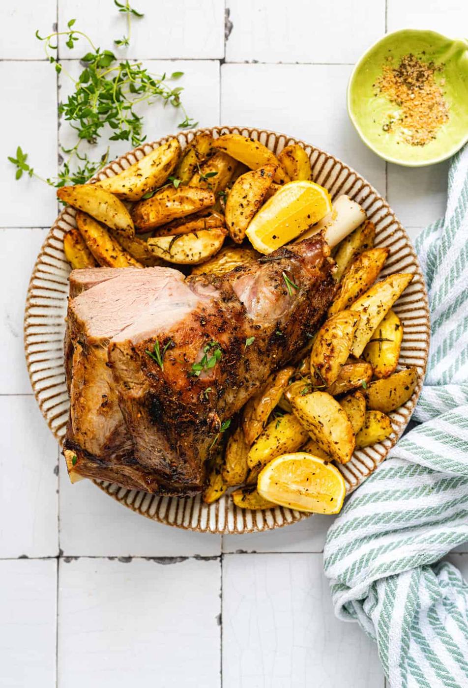 An Air Fryer Roast Lamb leg sits on a plate, garnished with herbs and lemon wedges, served alongside seasoned potato wedges. A bowl of seasoning and a striped cloth napkin rest nearby on the tiled surface.