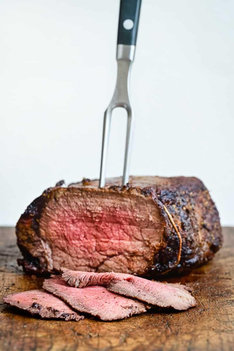 A juicy, medium-rare Air Fryer Roast Beef sits on a wooden cutting board, partially sliced with two pieces in front, and a meat fork inserted into the top.