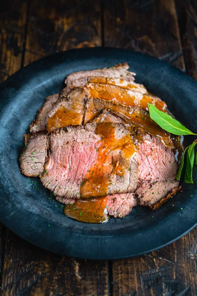 Slices of medium-rare Air Fryer Roast Beef topped with brown gravy are served on a dark plate, garnished with a green herb sprig, on a rustic wooden table.