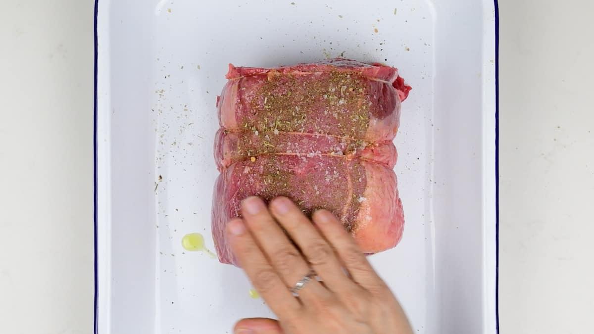 A hand rubs seasoning and oil onto a tied, raw beef roast placed in a white rectangular baking dish, prepping it perfectly for Air Fryer Roast Beef.