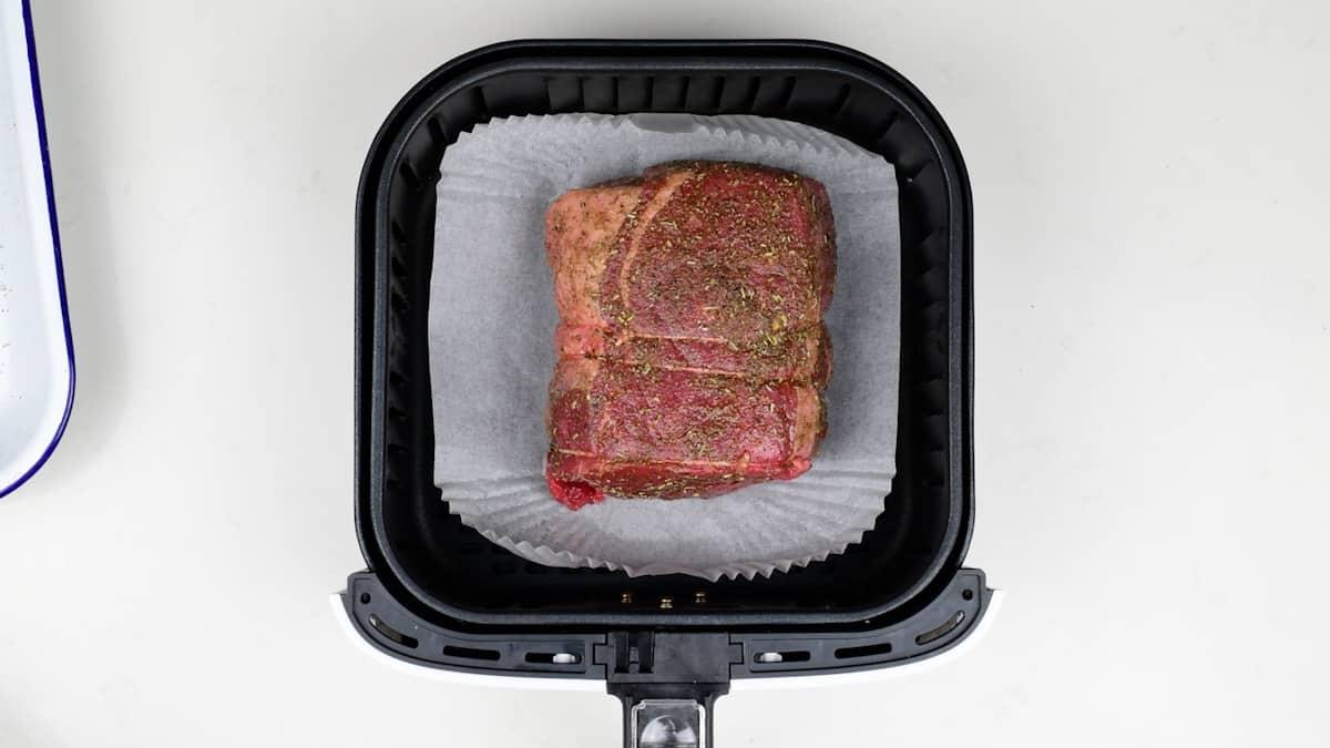 A seasoned Air Fryer Roast Beef sits on parchment paper inside the basket, ready to be cooked.