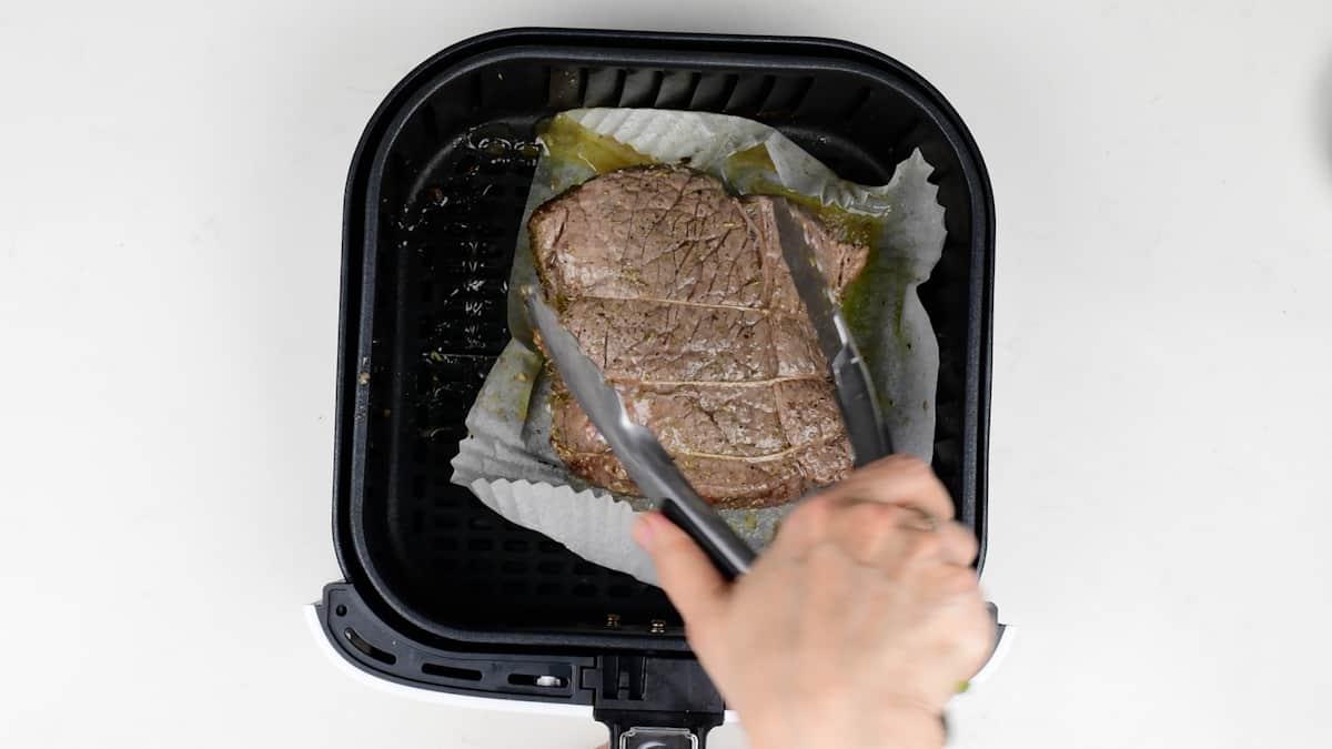 A hand holds tongs, lifting a cooked piece of Air Fryer Roast Beef resting on parchment paper inside an air fryer basket, viewed from above.
