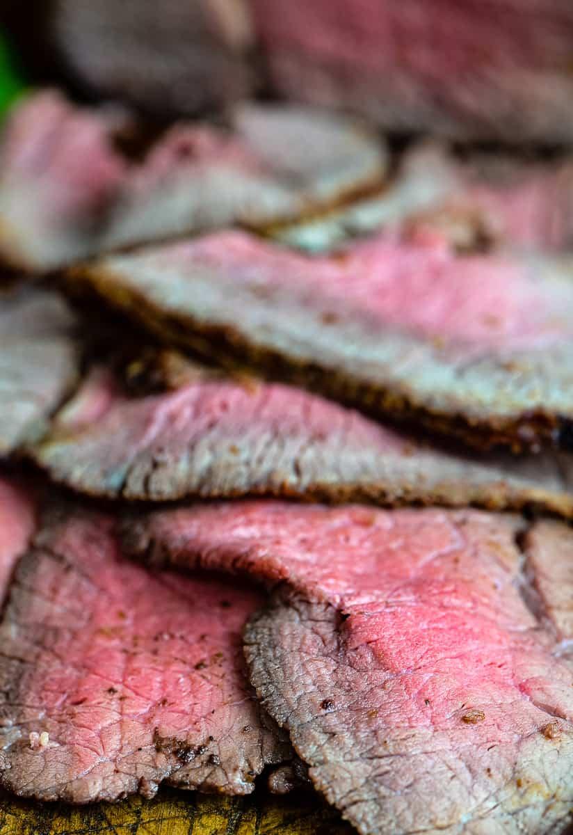 Close-up of several slices of Air Fryer Roast Beef, with a pink center and browned edges, stacked on a wooden cutting board.